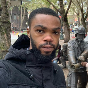 Picture of Bright Agbodzi, MS MPhil, in an outdoor plaza lined with trees, while another person interacts with a group of life‑sized firefighter statues arranged as if operating a fire hose